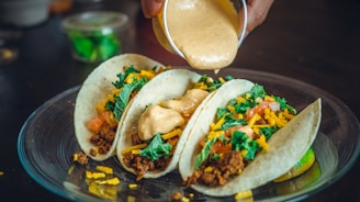 Three soft tacos filled with ground meat, chopped tomatoes, shredded cheddar cheese, and fresh greens, placed on a transparent plate. A hand is pouring a creamy sauce over the tacos. A small container of green herbs is visible in the background.
