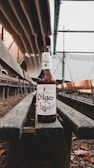 A beer bottle labeled 'Pilger' is positioned on a wooden bench within an industrial setting. The background includes metal structures and beams.
