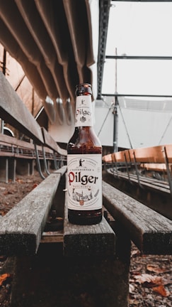 A beer bottle labeled 'Pilger' is positioned on a wooden bench within an industrial setting. The background includes metal structures and beams.