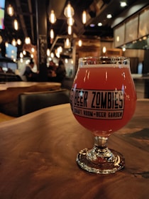 A glass of reddish-orange beer is placed on a wooden table in a cozy, dimly lit bar. The glass has a label that reads 'Beer Zombies Draft Room Beer Garden.' In the background, there are Edison-style hanging lights and a blurred view of people and bar interior.