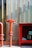 A fire hydrant and a fire hose reel box against a backdrop of large vertical panels. The hydrant is bright red, contrasting with the muted tones of the wall behind. The panels have a semi-transparent design, revealing faint silhouettes of plants. Sunlight casts shadows, adding depth to the scene.