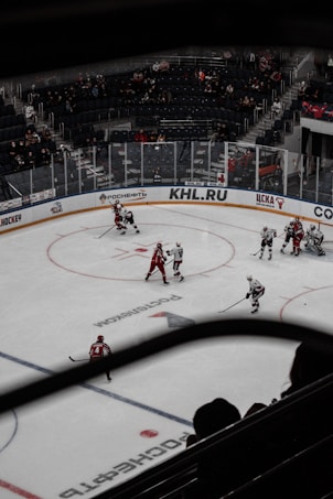 A hockey game is taking place in an indoor rink, with players dressed in red and white uniforms. The ice is marked with various logos, and there are spectators in the stands, though the seats are not completely filled.