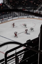 A hockey game is taking place in a large arena with players in red and white jerseys on the ice. The arena is partially filled with spectators. Advertising boards surround the rink with visible brands and logos.