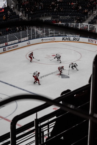 A hockey game is taking place in a large arena with players in red and white jerseys on the ice. The arena is partially filled with spectators. Advertising boards surround the rink with visible brands and logos.