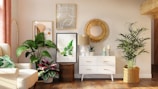A well-decorated living space featuring a white dresser with gold handles, topped with a lamp, two candles, and a small plant. Artwork is stylishly arranged on the wall, including a round wooden mirror and framed leaf prints. Large green plants are placed on either side of the dresser, complementing the warm and inviting ambiance of the room. A comfortable armchair with a green leaf-patterned cushion is positioned to the left, next to a floor lamp.