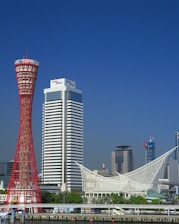 A vibrant cityscape featuring a distinctive red lattice tower next to a tall, modern hotel building. In the foreground is a waterfront area with lush greenery and a unique white architectural structure. Additional skyscrapers can be seen in the background under a clear blue sky.