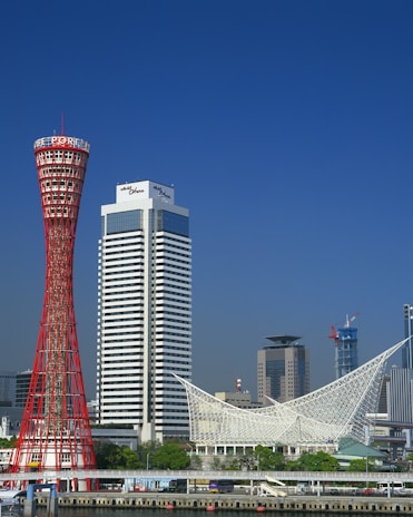 A vibrant cityscape featuring a distinctive red lattice tower next to a tall, modern hotel building. In the foreground is a waterfront area with lush greenery and a unique white architectural structure. Additional skyscrapers can be seen in the background under a clear blue sky.