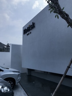 A minimalist building facade with a sign that reads 'Kotask', accompanied by a sleek modern car parked beside it. The building has a light gray finish and is situated near a small water feature. A tree is visible on the right side of the image, adding a natural element to the modern setting. The sky is slightly overcast.