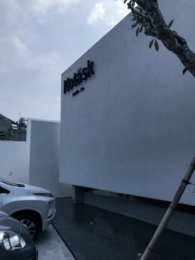 A minimalist building facade with a sign that reads 'Kotask', accompanied by a sleek modern car parked beside it. The building has a light gray finish and is situated near a small water feature. A tree is visible on the right side of the image, adding a natural element to the modern setting. The sky is slightly overcast.