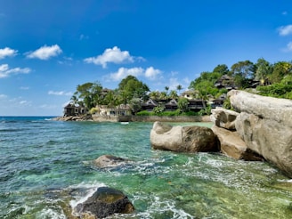 A scenic coastal landscape featuring clear turquoise waters lapping against large rocks in the foreground. Further back, a group of tropical-style villas nestled amongst lush green trees sits by the edge of the sea. The sky is bright blue with a few scattered white clouds.
