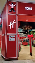 A toy store entrance with vibrant red walls and signage. Inside, shelves are filled with various toys and products. The entrance features whimsical decor, including cartoon character designs and colorful displays. Overhead lighting and decorative elements enhance the playful atmosphere.