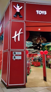 A toy store entrance with vibrant red walls and signage. Inside, shelves are filled with various toys and products. The entrance features whimsical decor, including cartoon character designs and colorful displays. Overhead lighting and decorative elements enhance the playful atmosphere.
