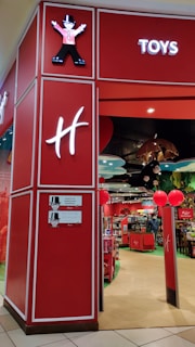 A toy store entrance with vibrant red walls and signage. Inside, shelves are filled with various toys and products. The entrance features whimsical decor, including cartoon character designs and colorful displays. Overhead lighting and decorative elements enhance the playful atmosphere.