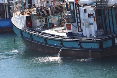 A sizable fishing boat is docked, featuring several life buoys, metal railings, and equipment on the deck. The vessel displays a mix of blue, turquoise, and white colors, with visible water being pumped out of the side. The name and registration are painted on the side of the boat.