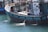 A sizable fishing boat is docked, featuring several life buoys, metal railings, and equipment on the deck. The vessel displays a mix of blue, turquoise, and white colors, with visible water being pumped out of the side. The name and registration are painted on the side of the boat.