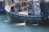 A sizable fishing boat is docked, featuring several life buoys, metal railings, and equipment on the deck. The vessel displays a mix of blue, turquoise, and white colors, with visible water being pumped out of the side. The name and registration are painted on the side of the boat.