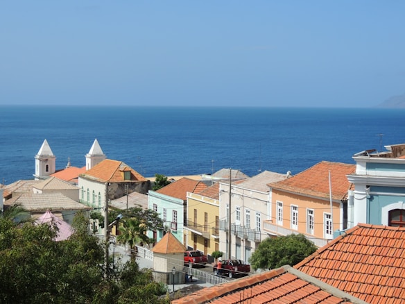 Colorful colonial-style buildings with red-tiled roofs are nestled along a coastline, with a deep blue ocean stretching into the horizon under a clear sky. Lush greenery and palm trees add vibrancy to the scene, and two white steeples are prominently featured among the buildings.