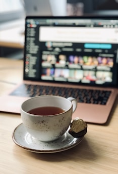 A ceramic cup filled with tea sits on a saucer, accompanied by a wrapped chocolate placed on the saucer. The background features a laptop with a blurred screen displaying colorful images, likely a video platform. The setting is on a wooden surface, suggesting a comfortable workspace or coffee table.