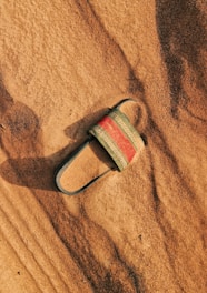 A single sandal lies partially buried in smooth, rippled sand. The sandal has a black sole and straps with a decorative red and white pattern across the top. Shadows and textures in the sand highlight the warm, earthy tones.