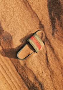 A single sandal lies partially buried in smooth, rippled sand. The sandal has a black sole and straps with a decorative red and white pattern across the top. Shadows and textures in the sand highlight the warm, earthy tones.