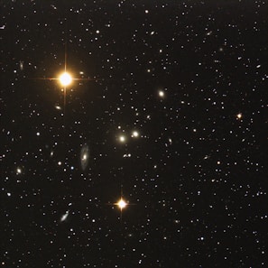 A vast expanse of deep space filled with numerous distant galaxies and stars, some of which are emitting a bright, golden light with noticeable diffraction spikes. The image captures countless smaller, faint specks of light scattered throughout the dark sky, suggesting the presence of far-off celestial bodies.