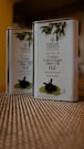 Two tin containers of Greek Tastes Premium Cretan Extra Virgin Olive Oil are placed on a textured surface. The design on the front includes olive branches and details about the product, including a 0.2 acidity level and a note that it is first cold pressed. The background is slightly blurred, giving emphasis to the tins.
