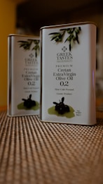 Two tin containers of Greek Tastes Premium Cretan Extra Virgin Olive Oil are placed on a textured surface. The design on the front includes olive branches and details about the product, including a 0.2 acidity level and a note that it is first cold pressed. The background is slightly blurred, giving emphasis to the tins.