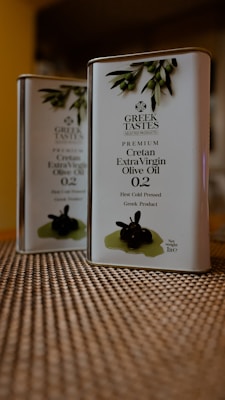 Two tin containers of Greek Tastes Premium Cretan Extra Virgin Olive Oil are placed on a textured surface. The design on the front includes olive branches and details about the product, including a 0.2 acidity level and a note that it is first cold pressed. The background is slightly blurred, giving emphasis to the tins.