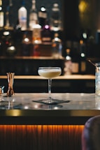 A cocktail glass filled with a creamy beverage sits atop a marble bar counter. The background is softly blurred, highlighting bottles of various spirits lined up on shelves, creating a warm and inviting atmosphere.