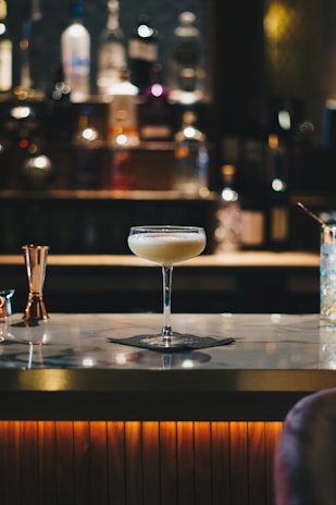 A cocktail glass filled with a creamy beverage sits atop a marble bar counter. The background is softly blurred, highlighting bottles of various spirits lined up on shelves, creating a warm and inviting atmosphere.