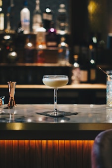 A cocktail glass filled with a creamy beverage sits atop a marble bar counter. The background is softly blurred, highlighting bottles of various spirits lined up on shelves, creating a warm and inviting atmosphere.