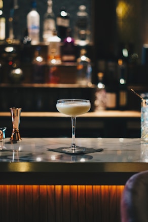 A cocktail glass filled with a creamy beverage sits atop a marble bar counter. The background is softly blurred, highlighting bottles of various spirits lined up on shelves, creating a warm and inviting atmosphere.
