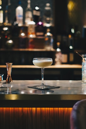 A cocktail glass filled with a creamy beverage sits atop a marble bar counter. The background is softly blurred, highlighting bottles of various spirits lined up on shelves, creating a warm and inviting atmosphere.