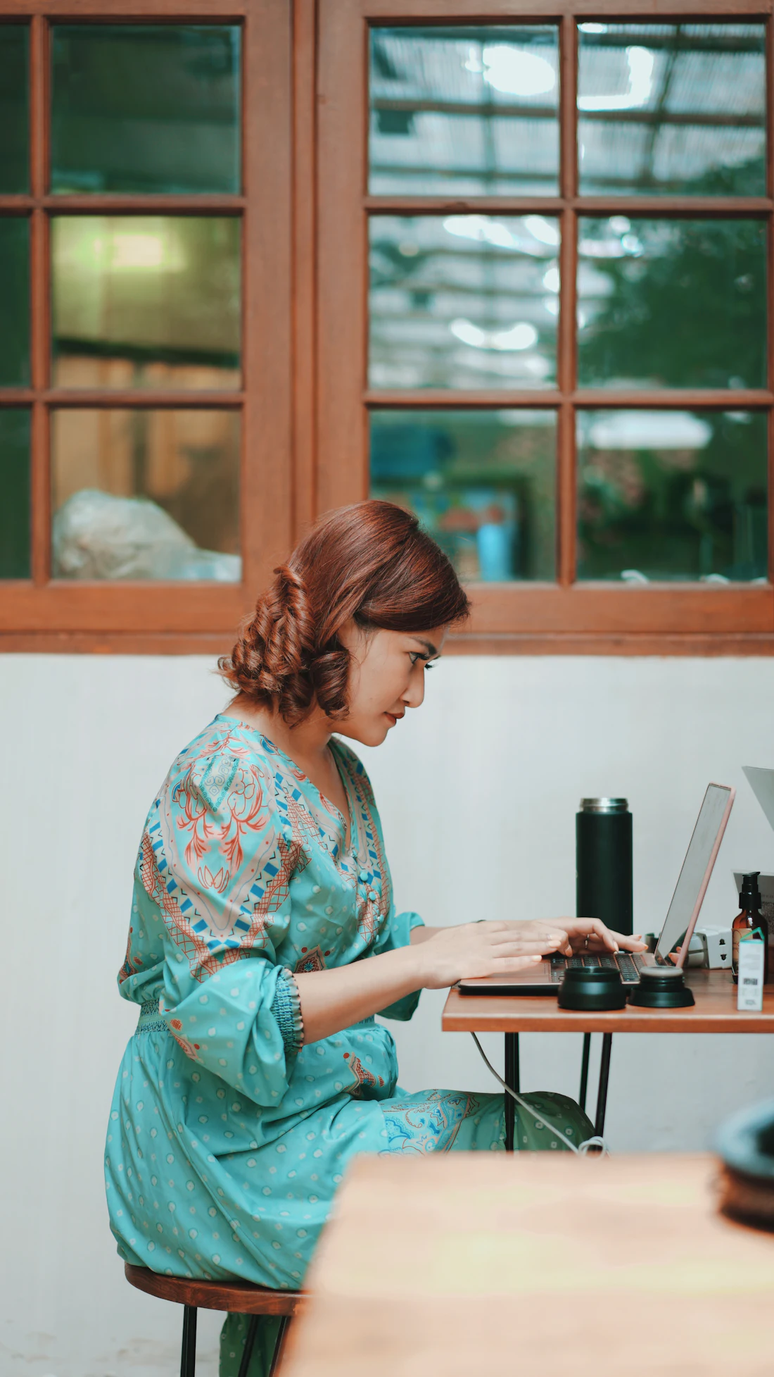 A woman in a batik dress typing on a laptop at a small table by a wooden window