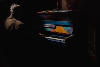 A person with gray hair, dressed in a brown checkered coat, is playing a brightly colored piano, featuring geometric patterns in shades of blue, yellow, and orange, amidst a dark and shadowy setting.