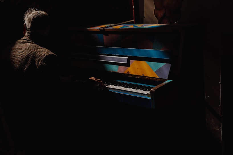 A person with gray hair, dressed in a brown checkered coat, is playing a brightly colored piano, featuring geometric patterns in shades of blue, yellow, and orange, amidst a dark and shadowy setting.