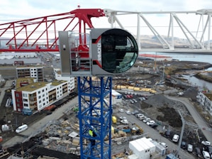 A large construction crane with a modern cabin is positioned above a developing construction site. The crane is red and white with a blue supporting structure. Below, there are numerous construction materials, vehicles, and unfinished buildings. Residential buildings with varying colors are visible in the background. The area appears industrial with a mix of urban development and natural scenery in the distance.