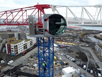 A large construction crane with a modern cabin is positioned above a developing construction site. The crane is red and white with a blue supporting structure. Below, there are numerous construction materials, vehicles, and unfinished buildings. Residential buildings with varying colors are visible in the background. The area appears industrial with a mix of urban development and natural scenery in the distance.