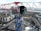 A large construction crane with a modern cabin is positioned above a developing construction site. The crane is red and white with a blue supporting structure. Below, there are numerous construction materials, vehicles, and unfinished buildings. Residential buildings with varying colors are visible in the background. The area appears industrial with a mix of urban development and natural scenery in the distance.