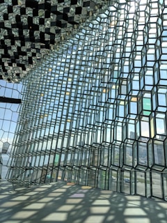 A large modern architectural structure features a geometrically intricate glass facade that captures and reflects light, creating a dynamic interplay of shadows and transparency. The outside view shows an urban landscape with buildings and a glimpse of water, contributing to a sense of openness and connection to the environment.