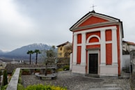 A quaint red and white chapel stands prominently with a backdrop of mountains. It is surrounded by a gravel courtyard with a stone bench and a small tree in a planter. The scene includes a few palm trees and additional rustic buildings with tiled roofs.