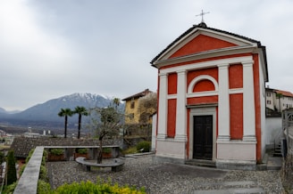 A quaint red and white chapel stands prominently with a backdrop of mountains. It is surrounded by a gravel courtyard with a stone bench and a small tree in a planter. The scene includes a few palm trees and additional rustic buildings with tiled roofs.