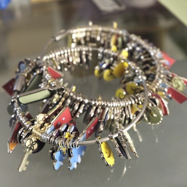 A collection of colorful keychains and small decorative trinkets attached to a circular metal band. The items include small keys, abstract shapes, and various metallic and painted charms in bright colors like red, yellow, and blue. The arrangement creates a visually striking mix of textures and forms.