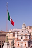 A large Italian flag waves prominently on a tall flagpole with the backdrop of classic European architecture. A domed building with ornate details stands out against a clear blue sky. Statues are visible on elevated structures, adding to the historic and cultural ambiance.