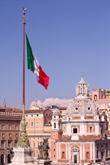 A large Italian flag waves prominently on a tall flagpole with the backdrop of classic European architecture. A domed building with ornate details stands out against a clear blue sky. Statues are visible on elevated structures, adding to the historic and cultural ambiance.