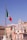 A large Italian flag waves prominently on a tall flagpole with the backdrop of classic European architecture. A domed building with ornate details stands out against a clear blue sky. Statues are visible on elevated structures, adding to the historic and cultural ambiance.