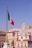 A large Italian flag waves prominently on a tall flagpole with the backdrop of classic European architecture. A domed building with ornate details stands out against a clear blue sky. Statues are visible on elevated structures, adding to the historic and cultural ambiance.