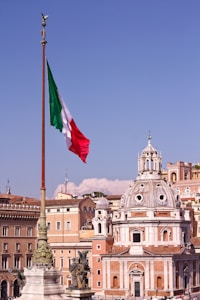 A large Italian flag waves prominently on a tall flagpole with the backdrop of classic European architecture. A domed building with ornate details stands out against a clear blue sky. Statues are visible on elevated structures, adding to the historic and cultural ambiance.