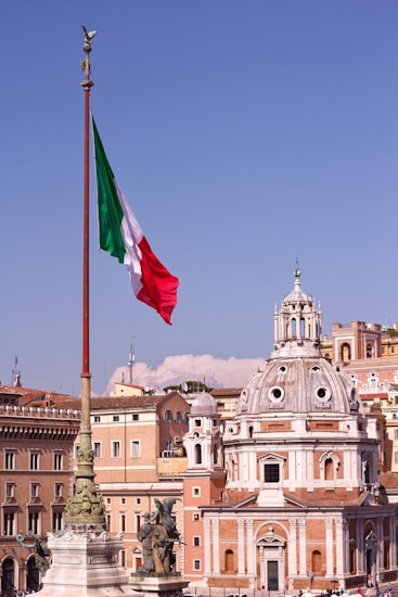 A large Italian flag waves prominently on a tall flagpole with the backdrop of classic European architecture. A domed building with ornate details stands out against a clear blue sky. Statues are visible on elevated structures, adding to the historic and cultural ambiance.