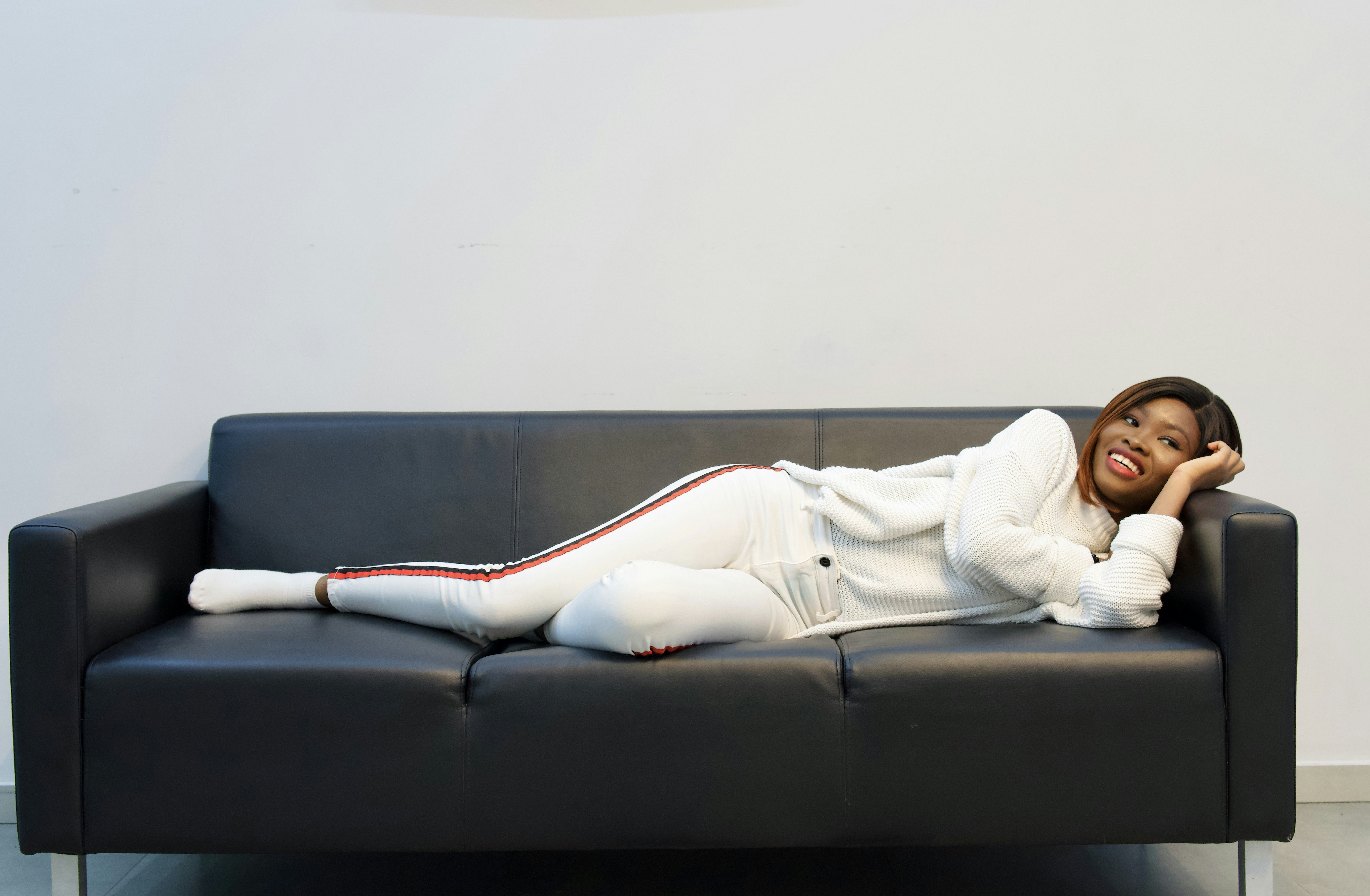 woman in white long sleeve shirt lying on black couch, Smiling Black girl lying on a black sofa and watching tv